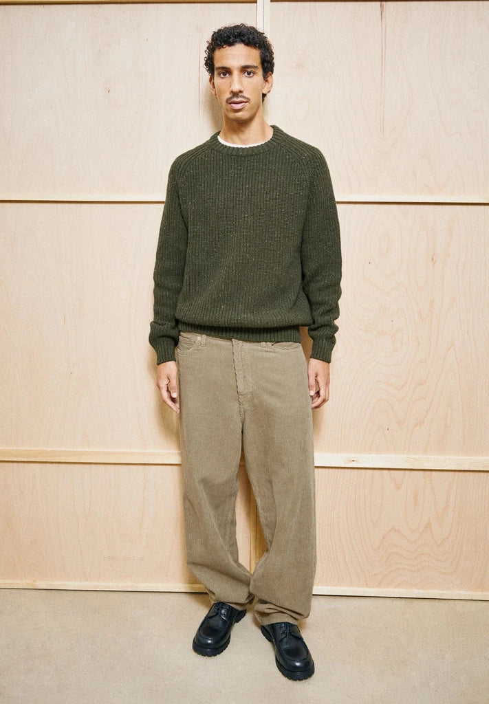 Man wearing the ARMEDANGELS Canada Kiaano Tweed Knit Sweater in Dark Earth Tweed over a white tee, standing in front of a plywood wall.