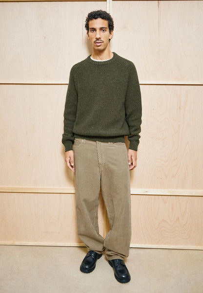 Man wearing the ARMEDANGELS Canada Kiaano Tweed Knit Sweater in Dark Earth Tweed over a white tee, standing in front of a plywood wall.