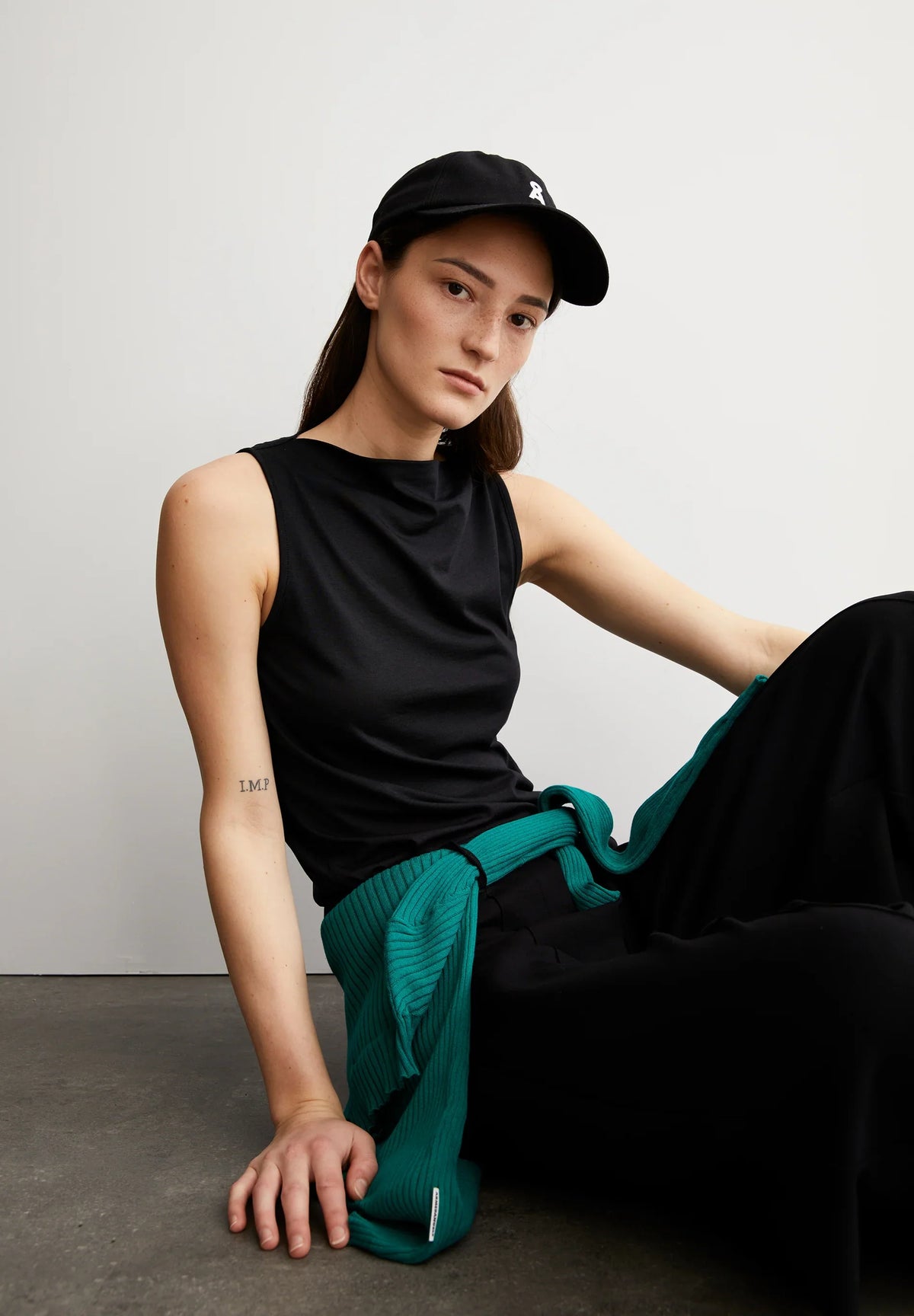 Woman in a black cap wearing the ARMEDANGELS Canada Laminiaa Sleeveless Top in black with black trousers and a green knit, seated on a concrete floor.