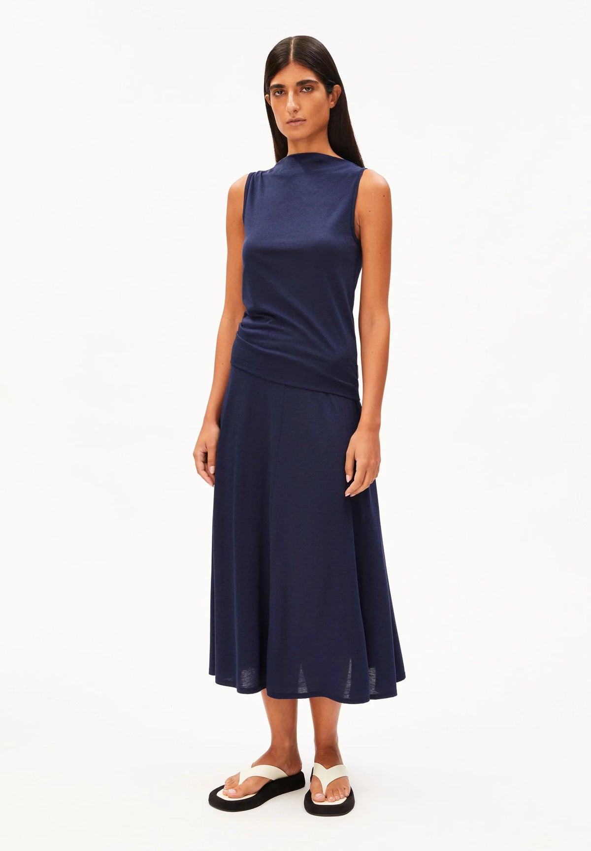 Full-length front view of a woman wearing the ARMEDANGELS Canada Laminiaa Sleeveless Top in tinted navy with a navy midi skirt and white sandals on a white background.