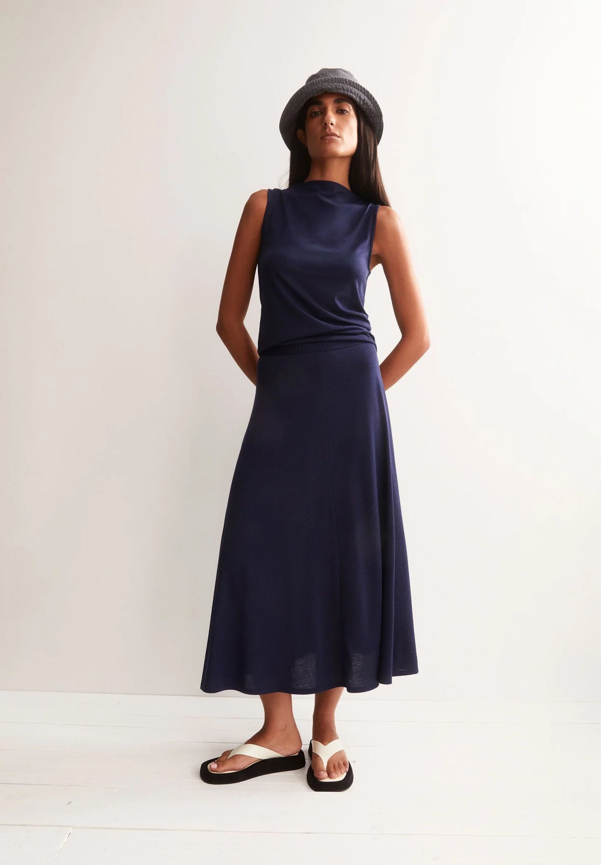 Woman wearing the ARMEDANGELS Canada Laminiaa Sleeveless Top in tinted navy with a navy midi skirt, grey bucket hat and white sandals, standing against a white wall.
