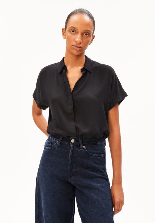 Woman wearing ARMEDANGELS Canada Larisaana blouse in black, tucked into dark blue jeans, against a white studio background.