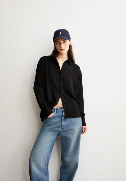 Woman wearing ARMEDANGELS Canada Larisaana Longsleeve Blouse in black with a navy cap and light-wash jeans, leaning against a light grey wall with one hand in her pocket.