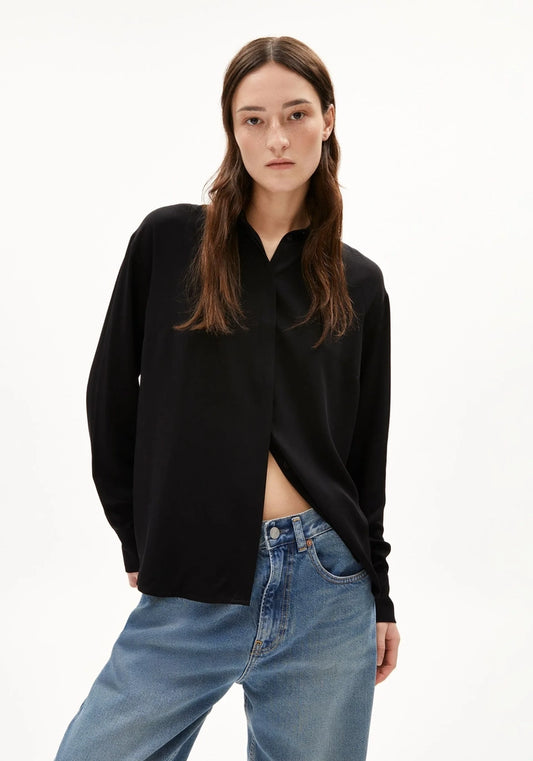 Woman wearing ARMEDANGELS Canada Larisaana Longsleeve Blouse in black, partly unbuttoned at the hem and paired with light-wash jeans, against a white background.