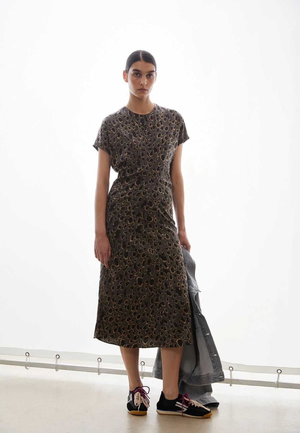 Woman wearing the ARMEDANGELS Canada Leo Bat Wing Midi Dress in leopard print with sneakers, holding a denim jacket in a bright white room.