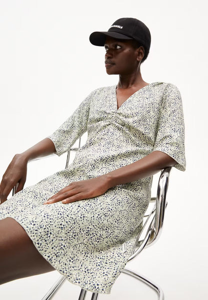 Woman in a black cap wearing the ARMEDANGELS Canada Lilaat Waterdrop Dress in seagreen, seated sideways on a chrome chair in a white studio.