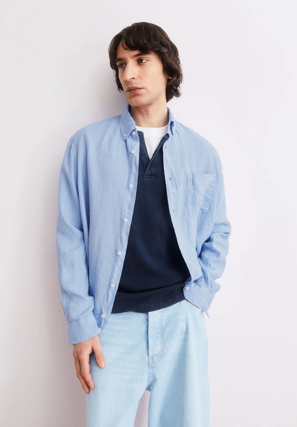 Man wearing the ARMEDANGELS Canada Linen Longsleeve Shirt in Wedgwood blue open over a navy knit and light jeans, leaning against a pale wall.