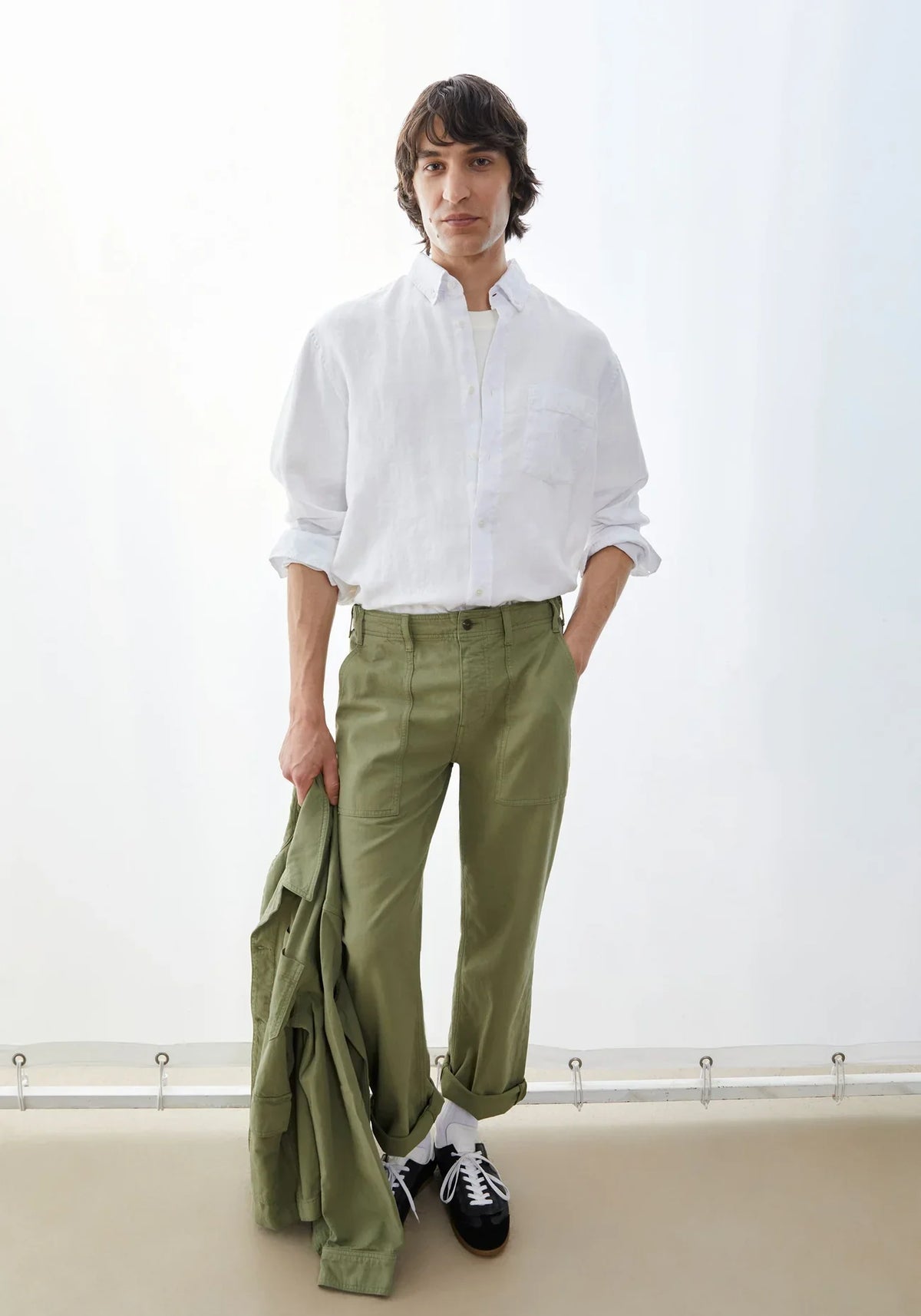 Man wearing the ARMEDANGELS Canada Linen Longsleeve Shirt in white with olive trousers, holding an olive jacket in a bright white room.