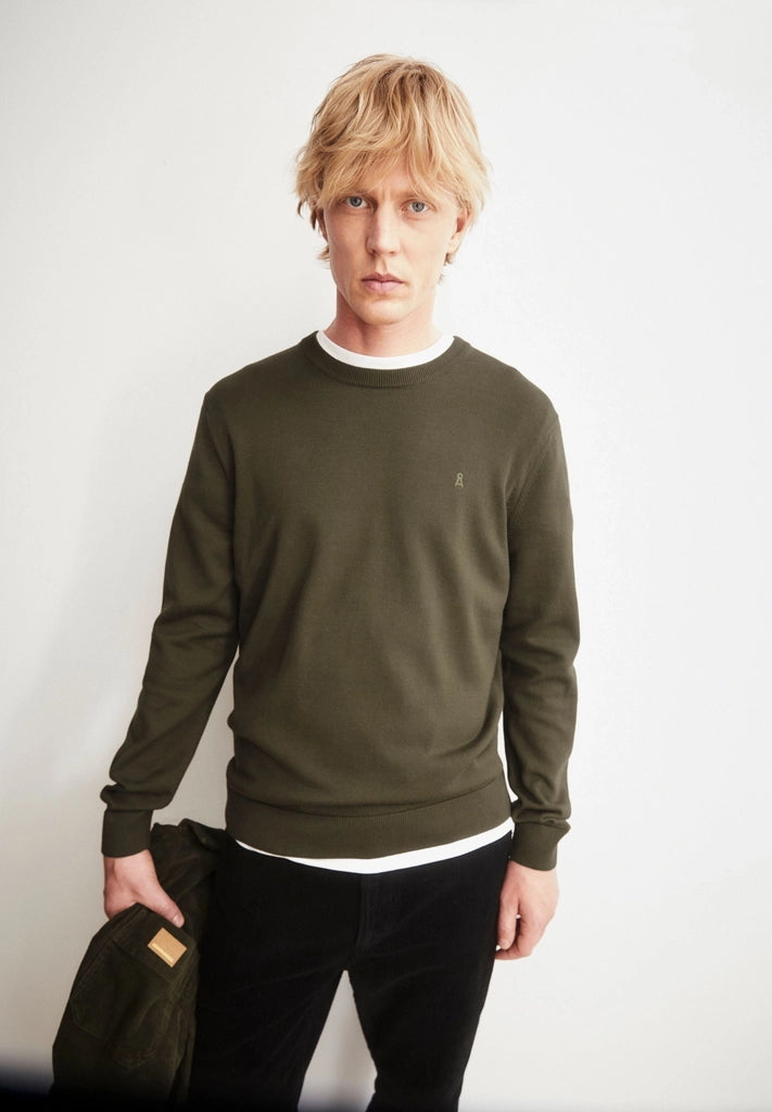 Man wearing the ARMEDANGELS Canada Loukaa Knit Sweater in dark earth over a white tee with black trousers, holding a dark jacket against a white wall.