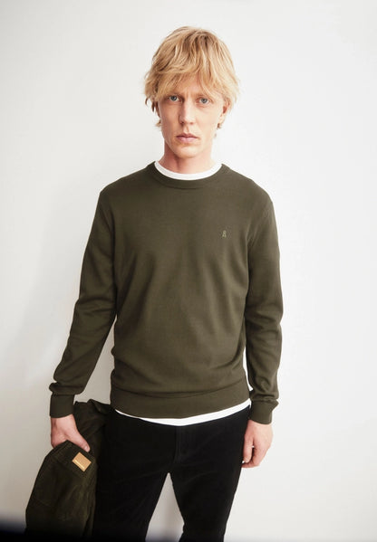 Man wearing the ARMEDANGELS Canada Loukaa Knit Sweater in dark earth over a white tee with black trousers, holding a dark jacket against a white wall.