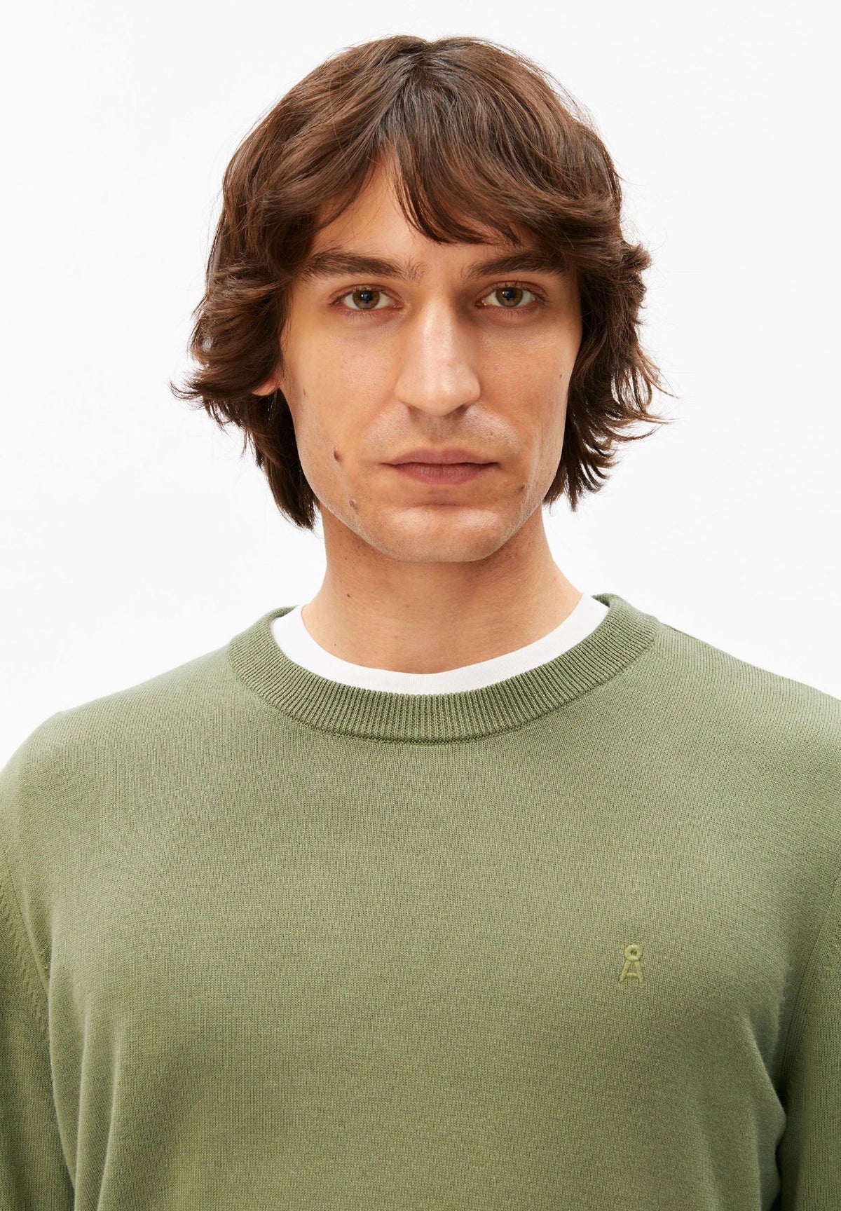 Close-up of the crew neck and tonal logo embroidery on the ARMEDANGELS Canada Loukaa Knit Sweater in fatigue green, worn by a man.