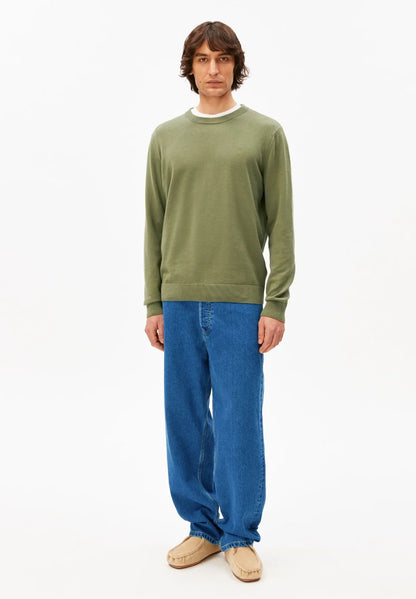 Full-length view of a man wearing the ARMEDANGELS Canada Loukaa Knit Sweater in fatigue green with blue jeans and suede shoes on a white background.