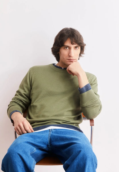 Man wearing the ARMEDANGELS Canada Loukaa Knit Sweater in fatigue green with blue jeans, seated on a chair against a pale pink wall.