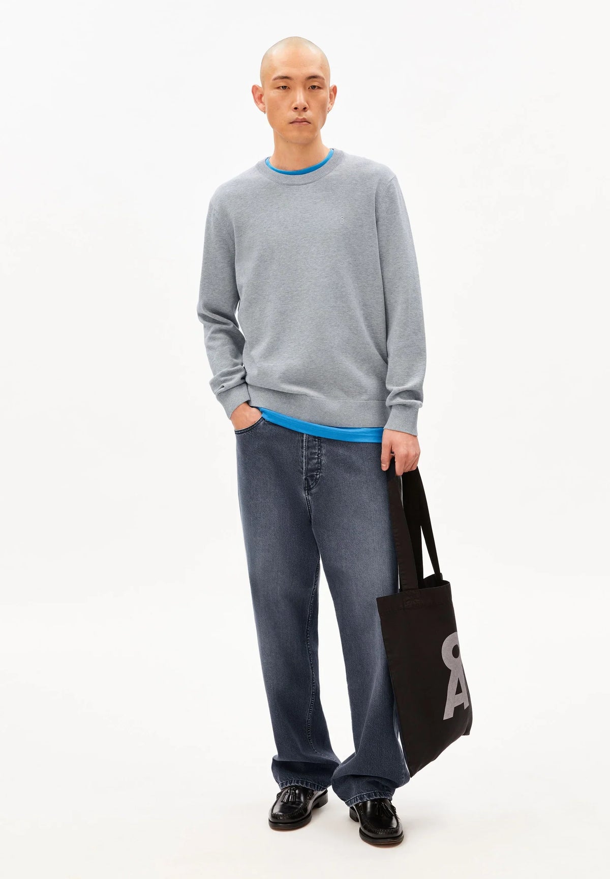 Full-length view of a man wearing the ARMEDANGELS Canada Loukaa Knit Sweater in silver melange with dark wide jeans, carrying a black tote bag, on a white background.