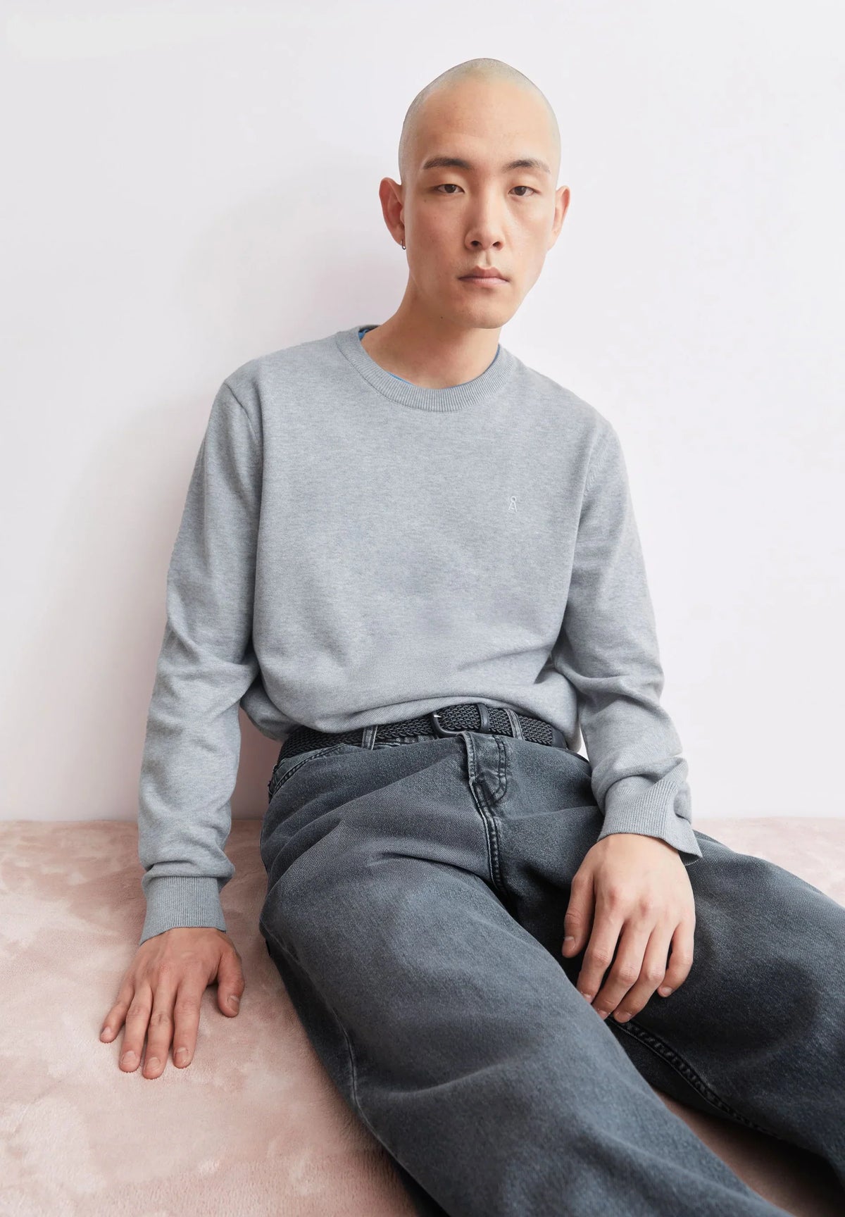 Man wearing the ARMEDANGELS Canada Loukaa Knit Sweater in silver melange with dark jeans, sitting on a pale pink floor against a pink wall.