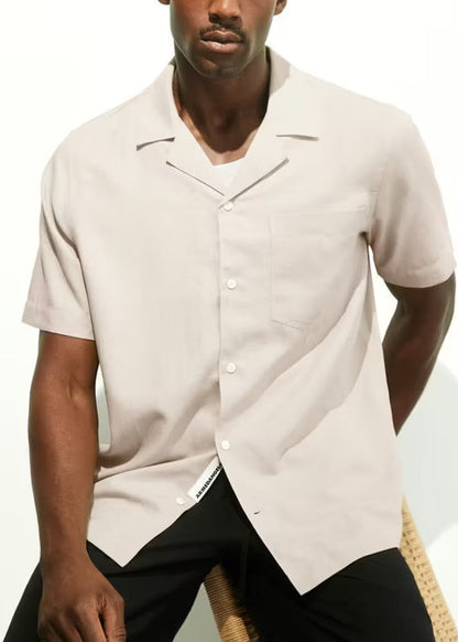 Man sitting on a wooden stool in the ARMEDANGELS Canada Lovaar Lino Shirt in sandstone, showing the camp collar, button placket and chest pocket, in a white studio.