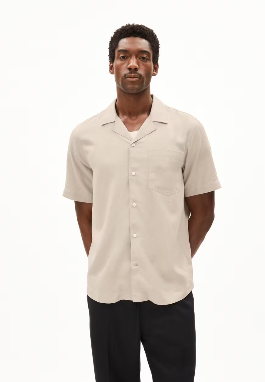 Man wearing the ARMEDANGELS Canada Lovaar Lino Shirt in sandstone, a relaxed short-sleeve shirt with a camp collar and chest pocket, with black trousers against a white studio background.