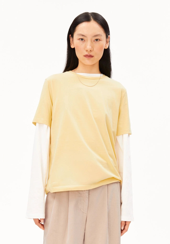 Woman wearing ARMEDANGELS Canada Lualaa T-Shirt in Golden Hay over a white long-sleeve top with beige trousers, front view against a white background.