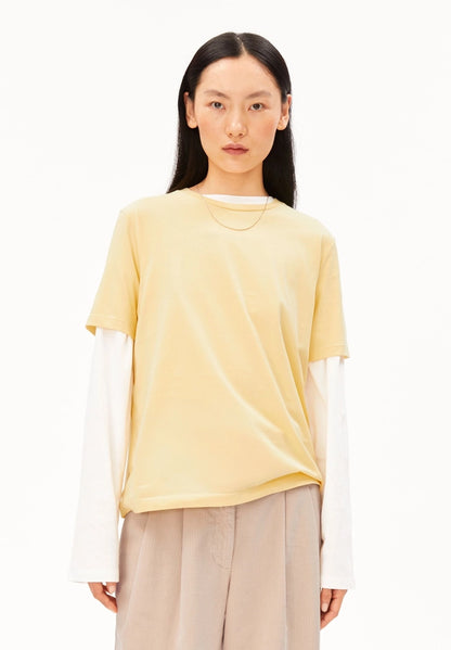 Woman wearing ARMEDANGELS Canada Lualaa T-Shirt in Golden Hay over a white long-sleeve top with beige trousers, front view against a white background.