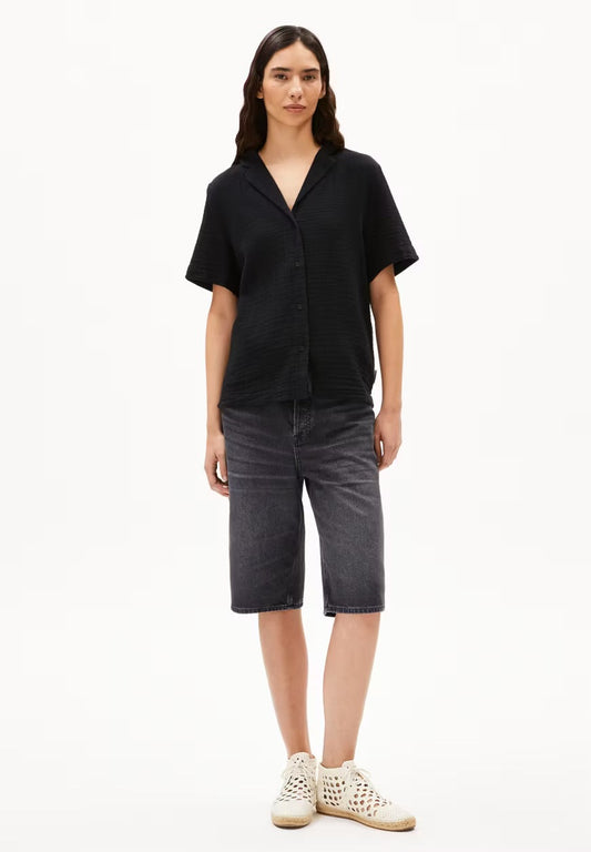 Woman wearing the ARMEDANGELS Canada Maaite Musselin Blouse in black, a relaxed short-sleeve muslin blouse with a revere collar, with grey denim shorts and white sneakers, against a white studio background.