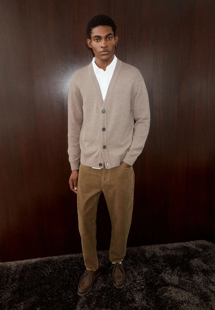 Man wearing ARMEDANGELS Canada Maakx Cord Trousers in Amberwood with a grey cardigan, standing in front of dark wood panelling.