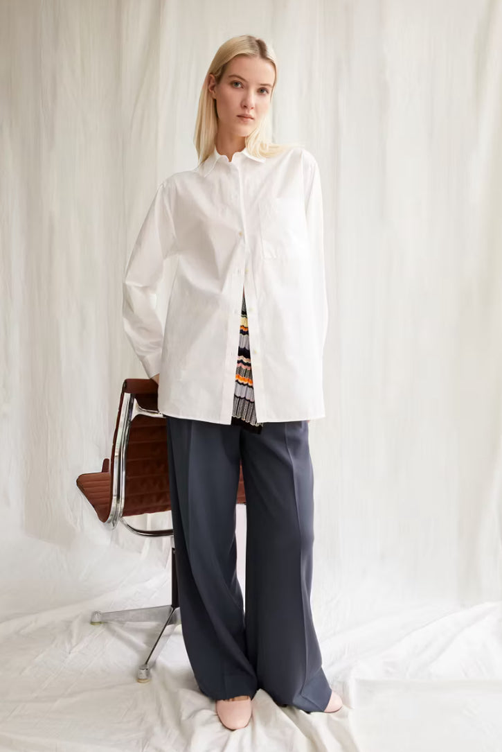 Woman wearing the ARMEDANGELS Canada Maalgaa Blouse in white with navy wide-leg trousers and a patterned skirt layer, standing by a chair in front of draped white fabric.