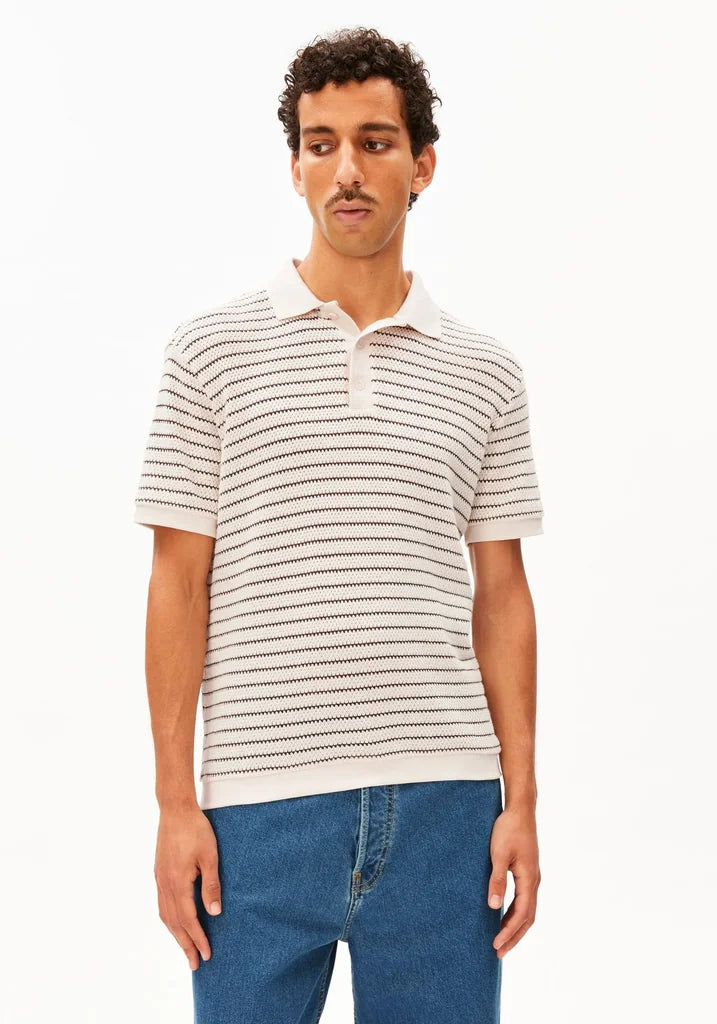 A man wearing a short-sleeve knit polo shirt with thin horizontal beige and black stripes, paired with blue jeans, against a plain white studio background.