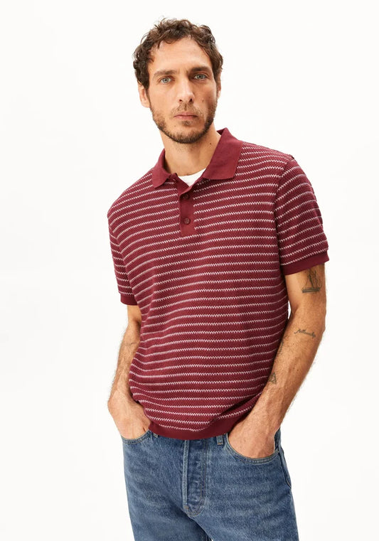 A man wearing a short-sleeve polo shirt with thin horizontal burgundy and white stripes, paired with blue jeans, standing against a plain white background.