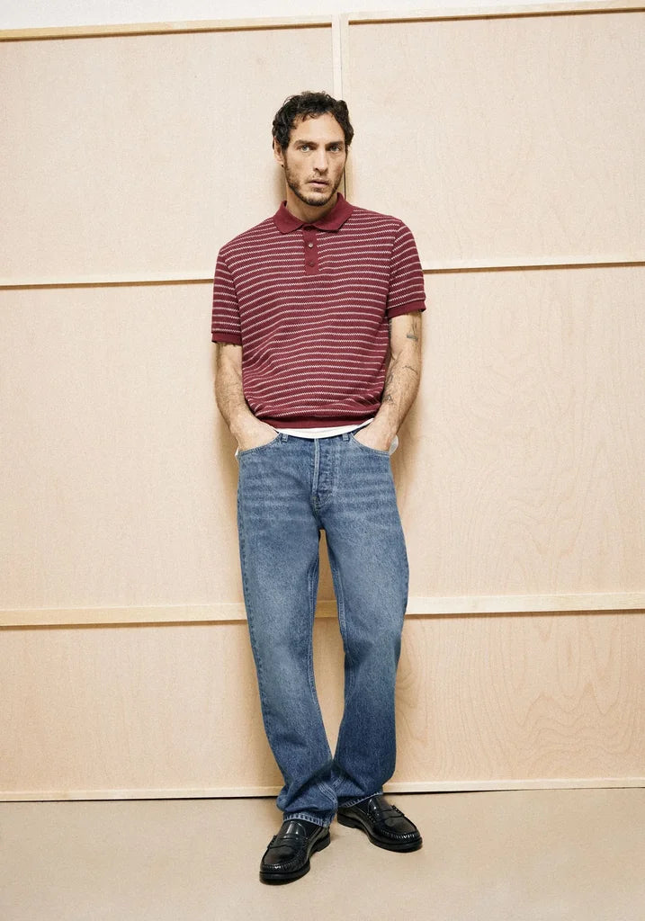 Man wearing a striped polo shirt and jeans standing against a wooden paneled wall.