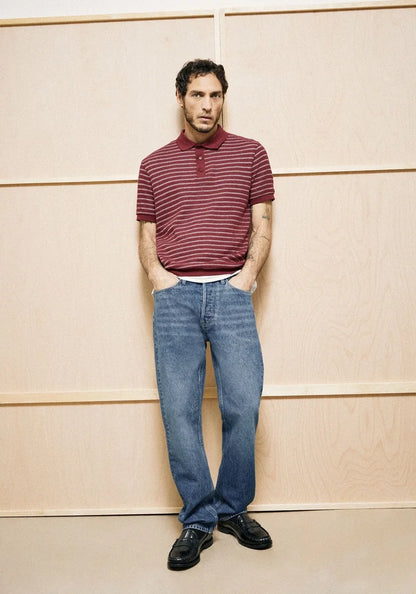 Man wearing a striped polo shirt and jeans standing against a wooden paneled wall.