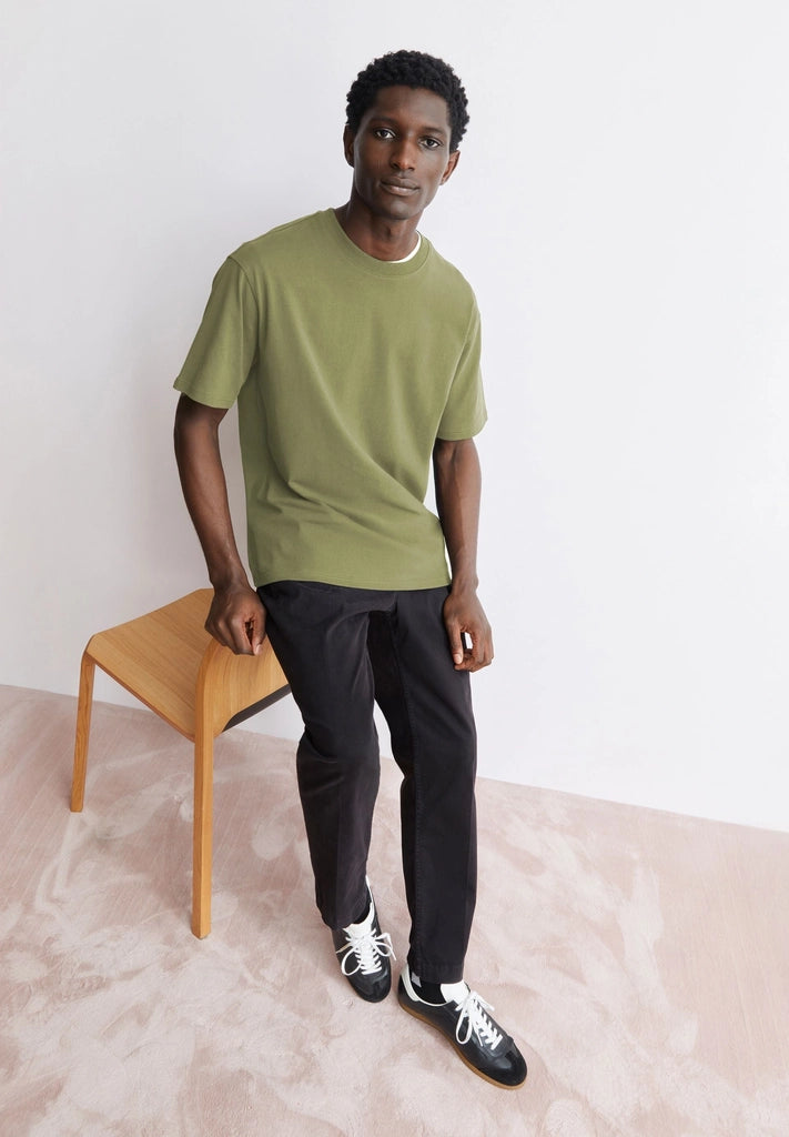 Man wearing the ARMEDANGELS Canada Maarkos Heavy Weight T-Shirt in fatigue green with black trousers, standing by a wooden stool in a bright room.