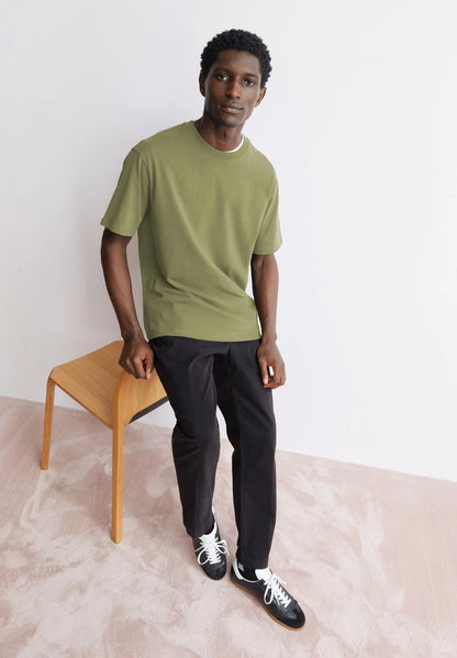 Man wearing the ARMEDANGELS Canada Maarkos Heavy Weight T-Shirt in fatigue green with black trousers, standing by a wooden stool in a bright room.