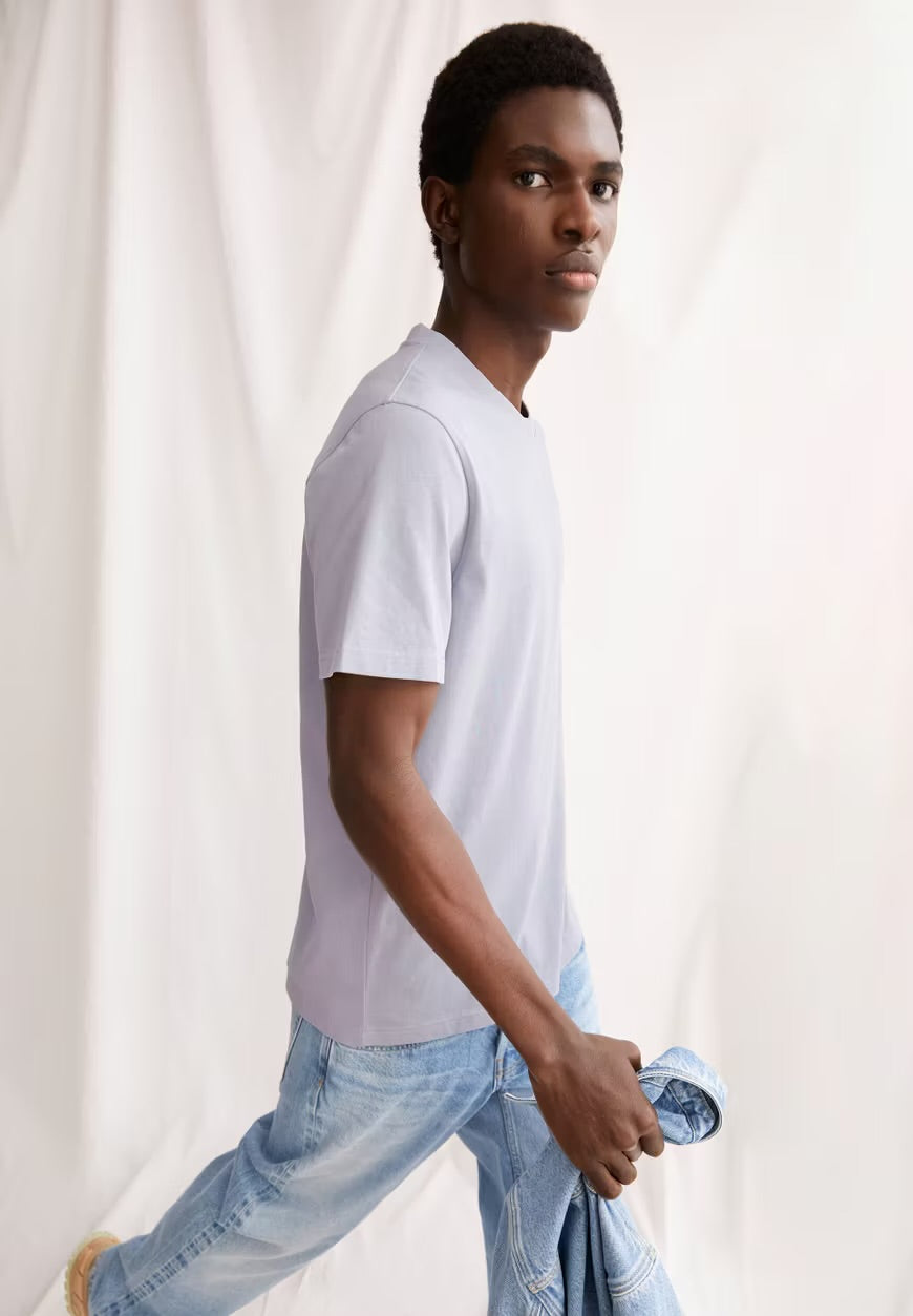 Man wearing the ARMEDANGELS Canada Maarkos Heavy Weight T-Shirt in misty morning with light jeans, mid-stride against a white curtain.