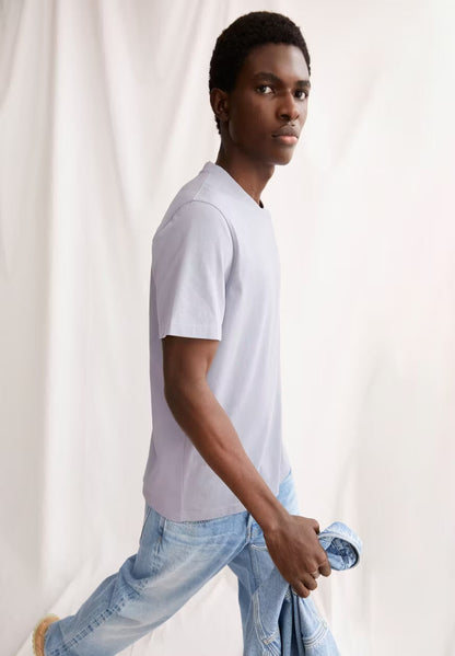Man wearing the ARMEDANGELS Canada Maarkos Heavy Weight T-Shirt in misty morning with light jeans, mid-stride against a white curtain.