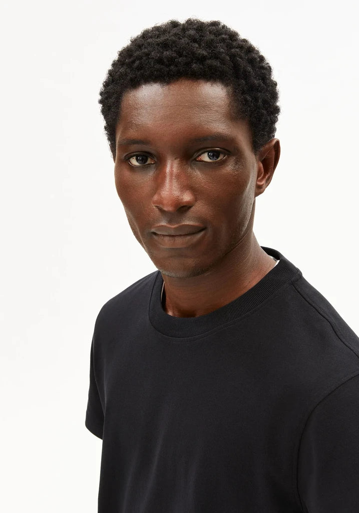 A close-up portrait of a man with short curly black hair wearing a solid black crew-neck t-shirt against a plain white background.