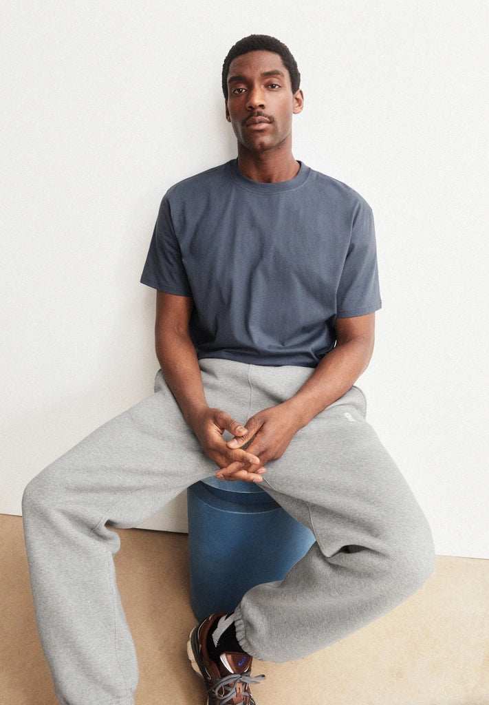 Man wearing the ARMEDANGELS Canada Maarkos Mid Weight T-Shirt in moonshadow with grey sweatpants, seated on a blue stool against a white wall.