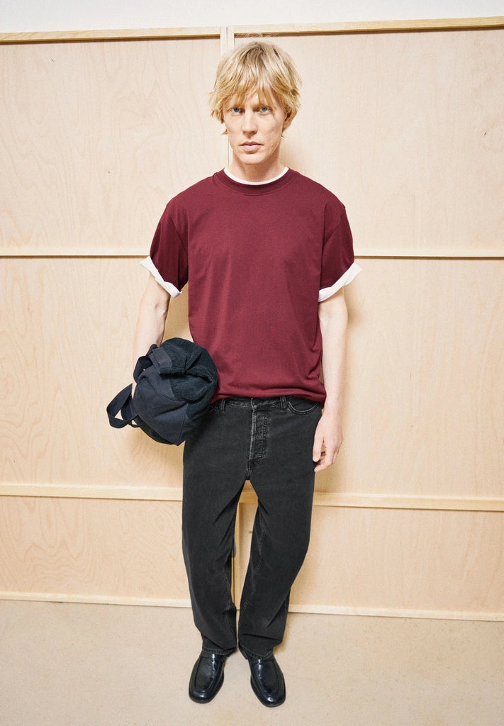 Man wearing the ARMEDANGELS Canada Maarkos Mid Weight T-Shirt in velvet red with black jeans, holding a navy jacket against a light wood wall.