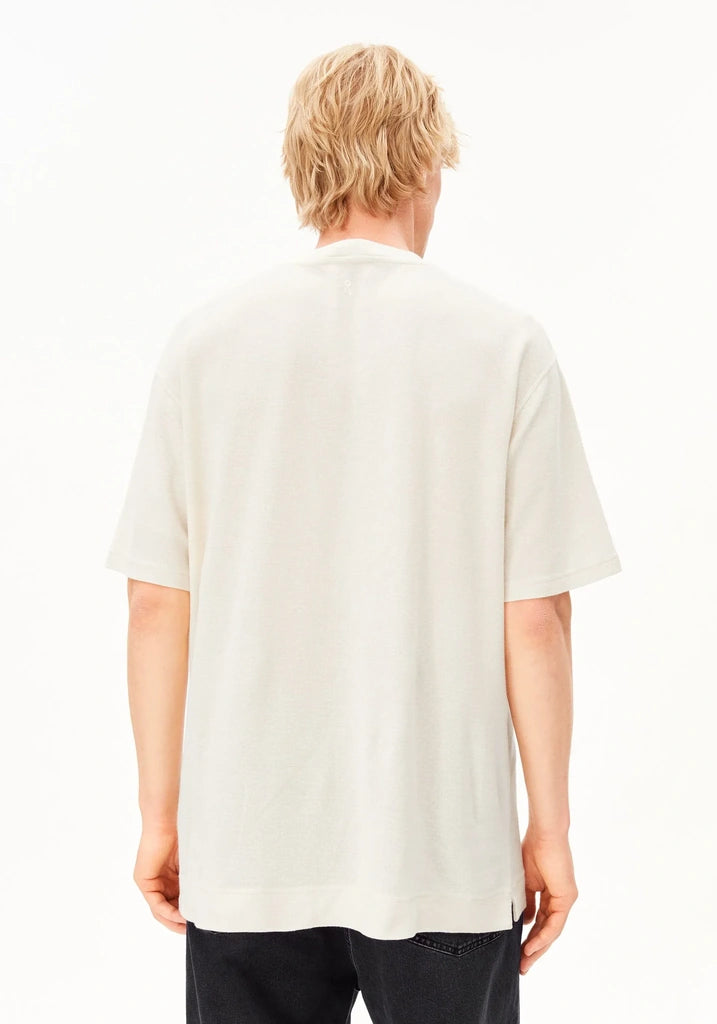 A man viewed from behind wearing an oversized off-white short-sleeve t-shirt and dark pants, set against a plain white studio background.