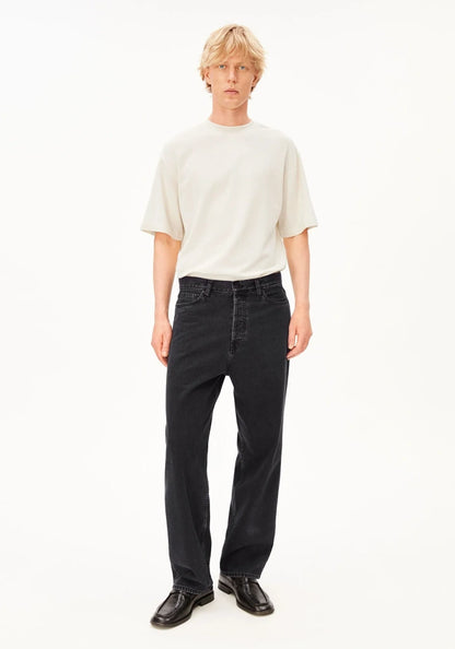A model stands against a plain white studio background wearing a cream-colored short-sleeve crewneck t-shirt, black wide-leg jeans, and black leather shoes.