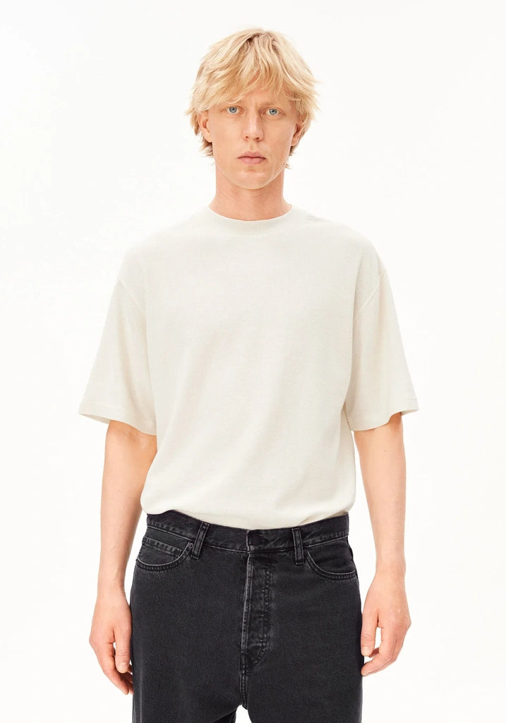 A blonde male model wearing a cream-colored short-sleeve crewneck t-shirt and dark wash jeans, standing against a plain white studio background.