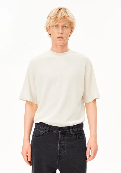 A blonde male model wearing a cream-colored short-sleeve crewneck t-shirt and dark wash jeans, standing against a plain white studio background.