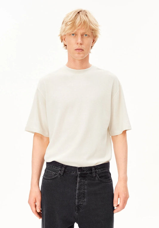 A blonde male model wearing a cream-colored short-sleeve crewneck t-shirt and dark wash jeans, standing against a plain white studio background.