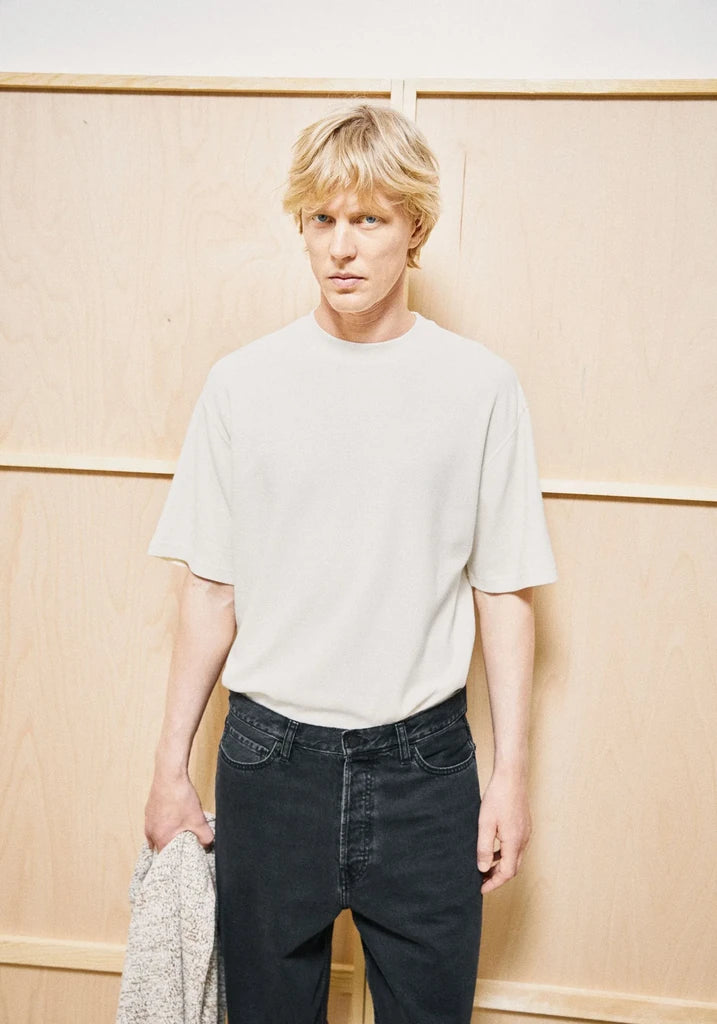 A blonde man wearing a light beige crewneck t-shirt and dark jeans, holding a gray knit sweater, standing against a light wood-paneled wall.