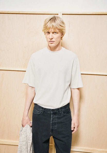 A blonde man wearing a light beige crewneck t-shirt and dark jeans, holding a gray knit sweater, standing against a light wood-paneled wall.