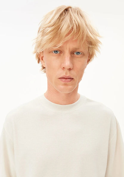 A close-up portrait of a person with short blonde hair and blue eyes, wearing a plain off-white crewneck sweater against a clean white background.