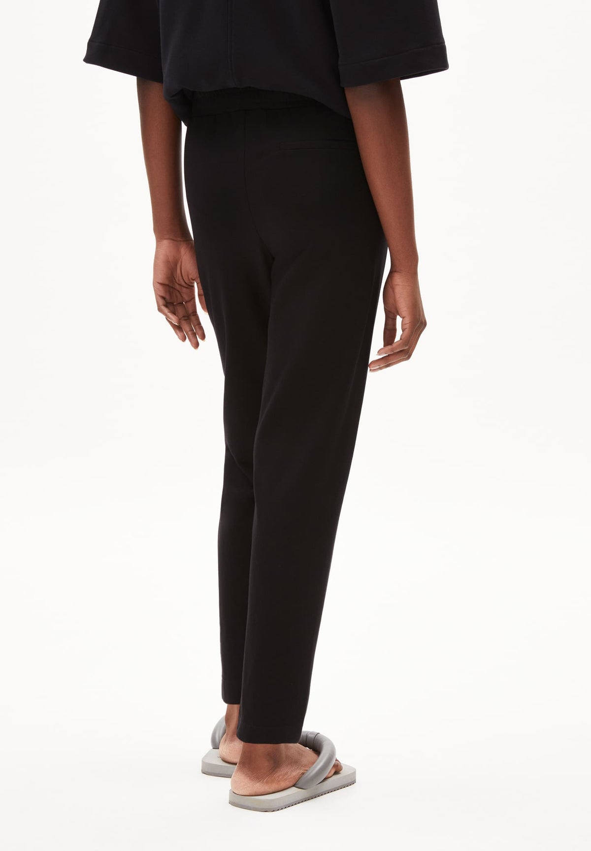 Back view of the ARMEDANGELS Canada Magdaalena Jersey Pant in black, showing the tapered leg, worn with grey slides against a white background.