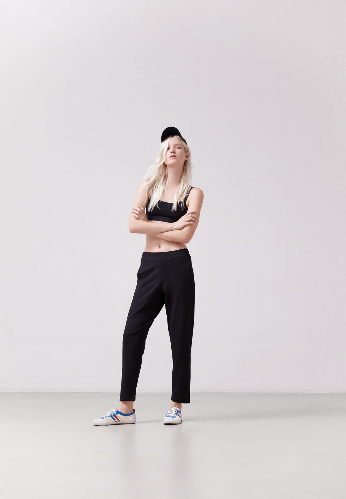 Woman wearing the ARMEDANGELS Canada Magdaalena Jersey Pant in black with a black bralette, cap and white sneakers, arms crossed, in a pale grey studio.
