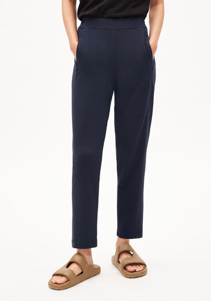 Front view of the ARMEDANGELS Canada Magdaalena Jersey Pant in night sky navy, hands in the side pockets, with tan sandals.