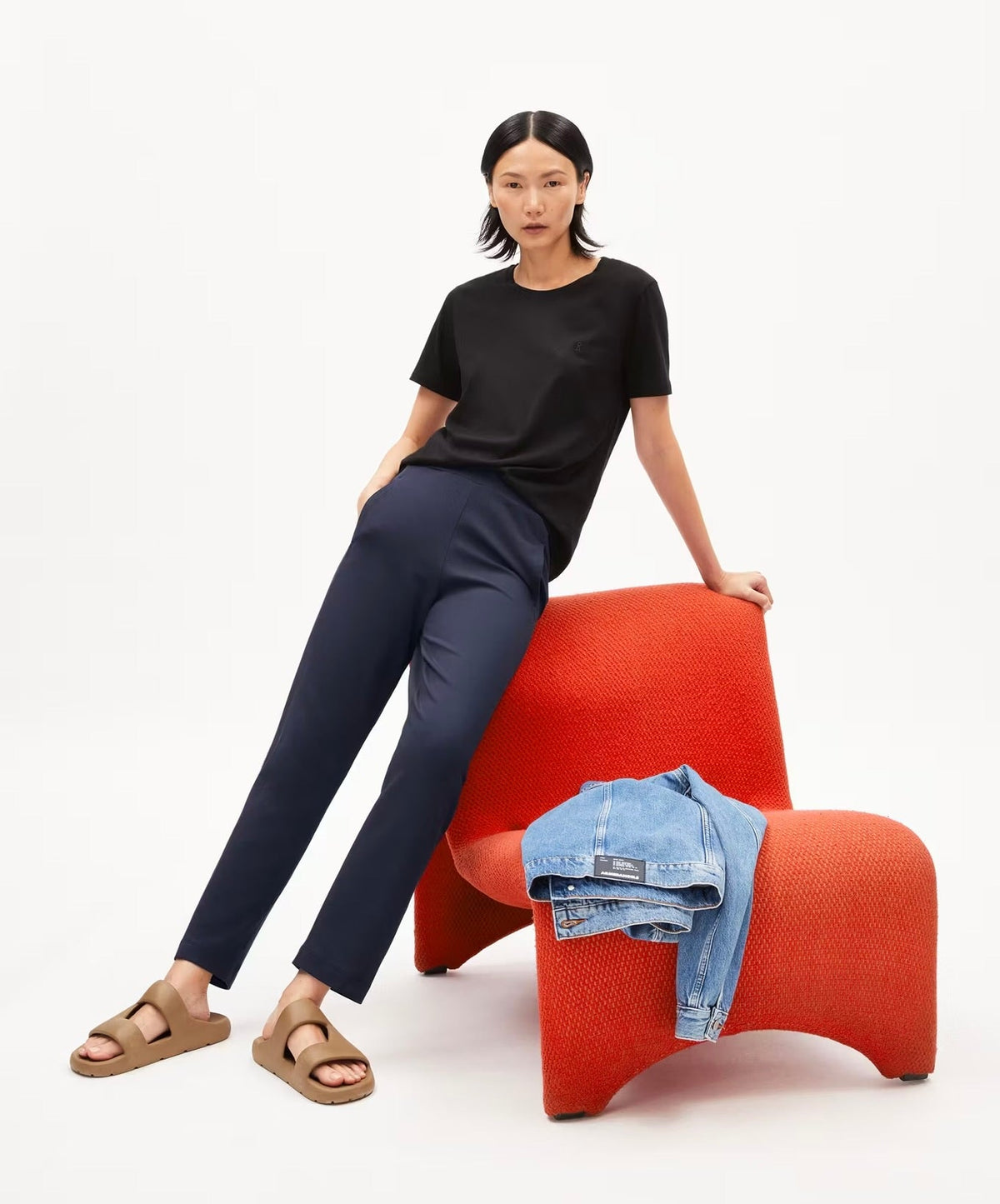 Woman leaning on an orange armchair in the ARMEDANGELS Canada Magdaalena Jersey Pant in night sky navy, with a black tee and tan sandals, in a white studio.