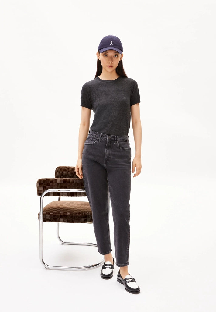Woman wearing ARMEDANGELS Canada Mairaa mom jeans in black standing beside a brown velvet chair against a white background.
